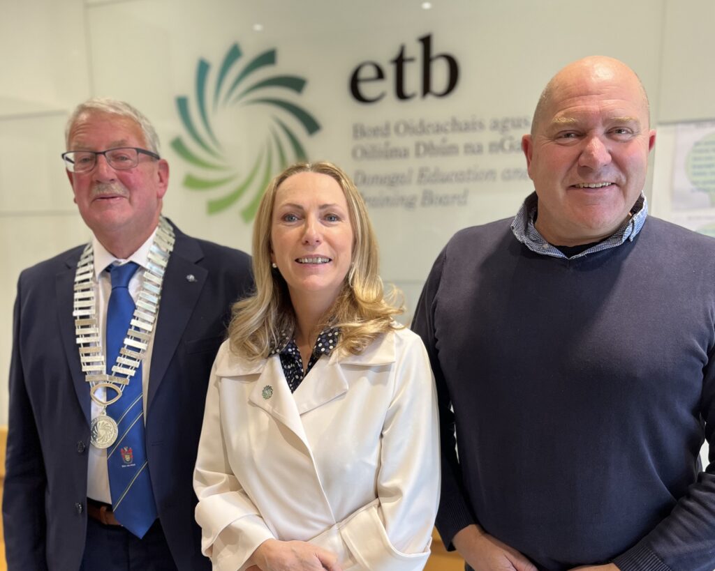 Three people standing side by side, one wearing a chain of office, in front of a wall with a logo on it.