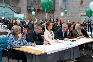 People sitting at a desk in a room of students.