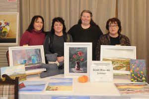 Four women standing behind a table with paintings on it.